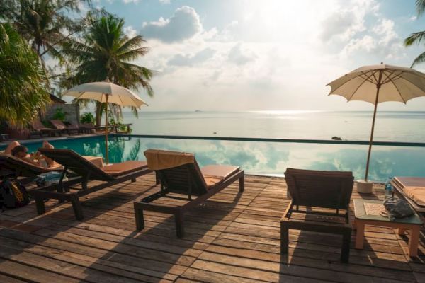 A serene poolside scene with lounge chairs, umbrellas, and palm trees overlooking a tranquil ocean under a partly cloudy sky.