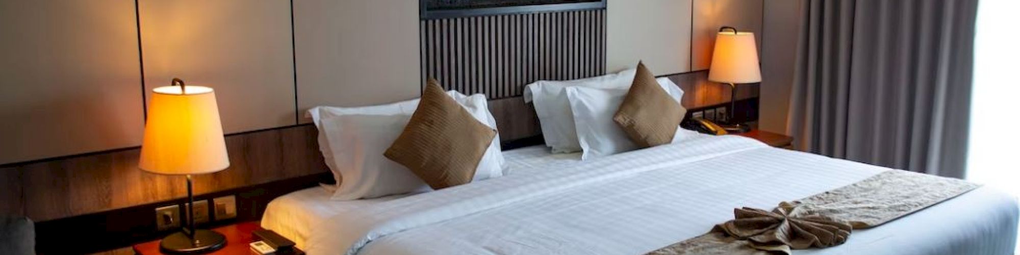 A cozy hotel room features a large bed with pillows, a warm-toned bedside lamp, and elegant decor, creating a relaxing atmosphere.