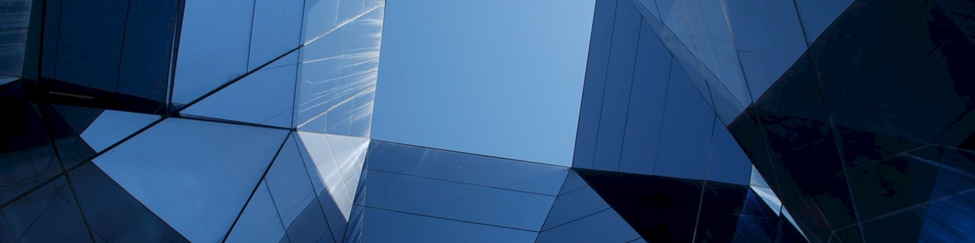 A geometric perspective looking up through modern glass architecture, reflecting a clear blue sky.