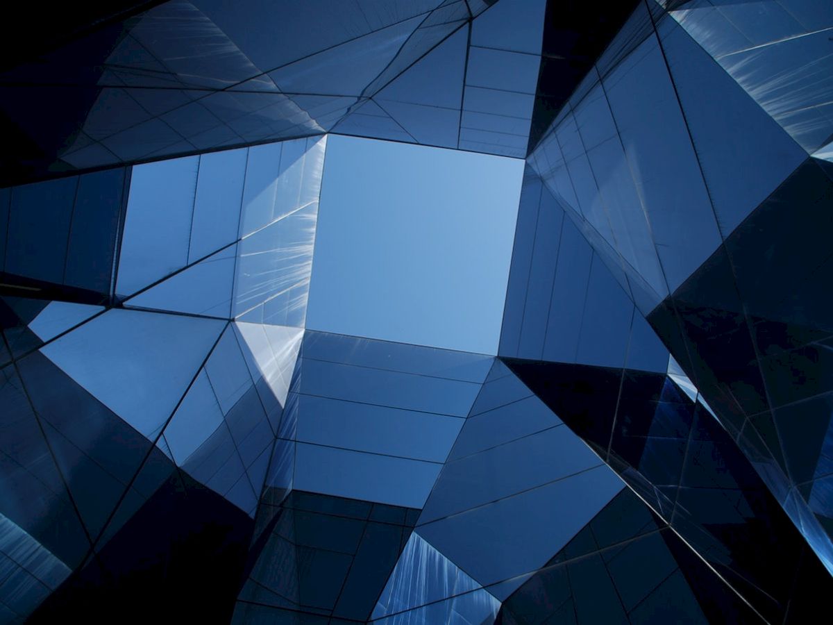 Abstract view from below of modern reflective glass buildings meeting at a geometric central point against a blue sky.