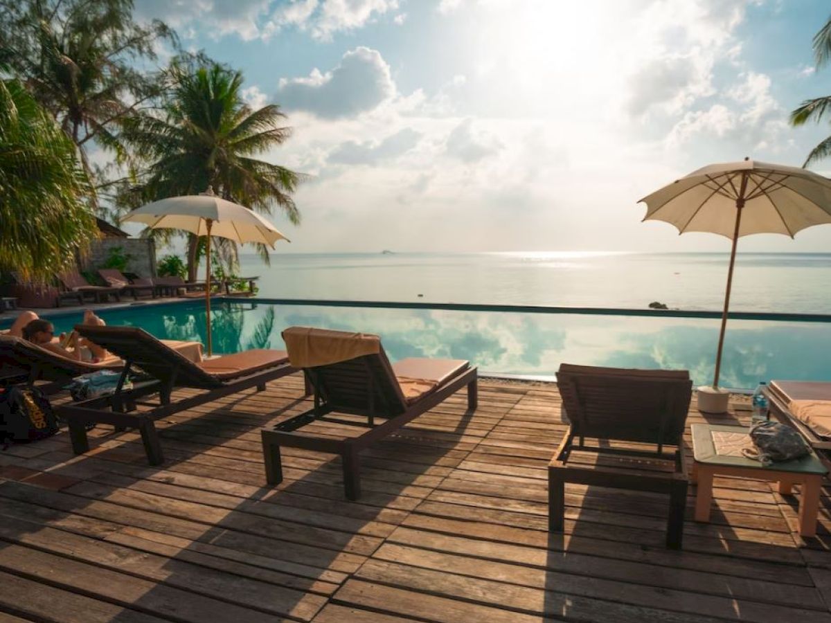 A tranquil poolside setting with lounge chairs, umbrellas, palm trees, and a view of the ocean under a partly cloudy sky.