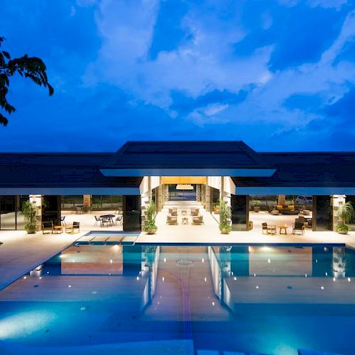 A modern building with large windows is overlooking a lit-up outdoor swimming pool at dusk, surrounded by outdoor seating.