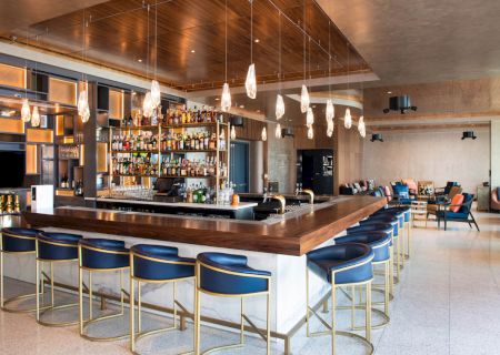 A stylish bar with a wooden counter, blue cushioned stools, hanging lights, and a fully stocked wall. Cozy seating in the background.