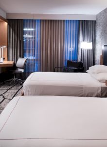 The image shows a modern hotel room with two beds, a wall-mounted TV, a desk, and large windows with curtains.