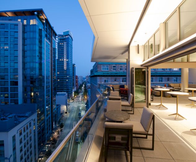 A modern balcony with tables and chairs overlooks a city street lined with tall buildings at dusk.
