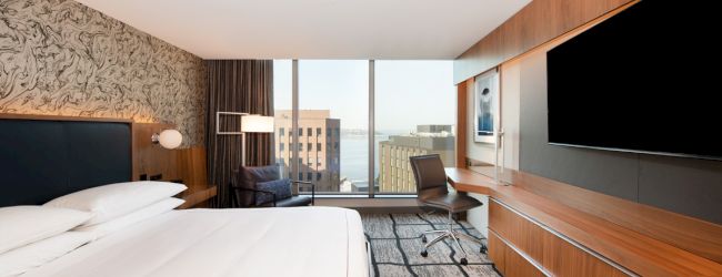 A modern hotel room with a bed, large TV, desk, and a window view of city buildings and water.