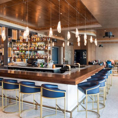 A modern bar with blue stools, wooden accents, pendant lights, and a stocked shelf. Cozy seating is visible in the background.