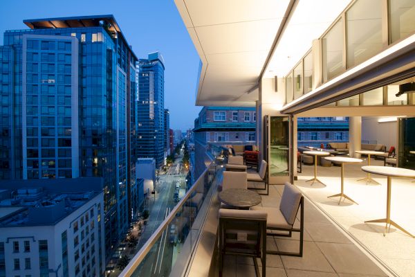 A modern cityscape at twilight, featuring tall buildings and a well-lit balcony with tables and chairs overlooking the street below.