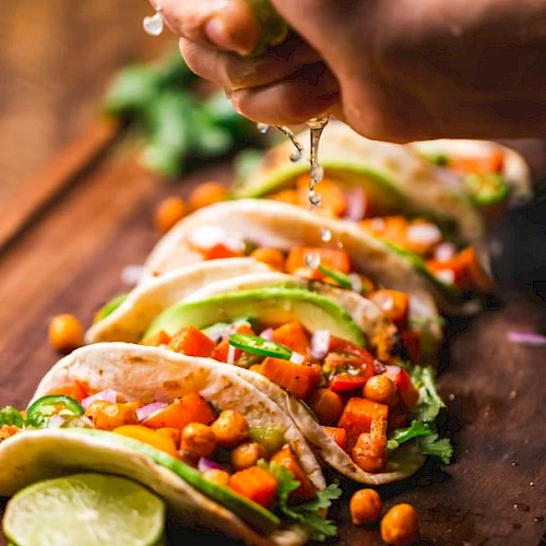 A person is squeezing lime over tacos filled with chickpeas, avocado, and vegetables arranged on a wooden board.