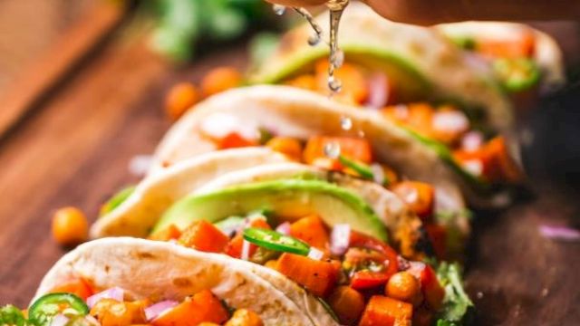 A person is squeezing lime over tacos filled with chickpeas, avocado, and vegetables arranged on a wooden board.
