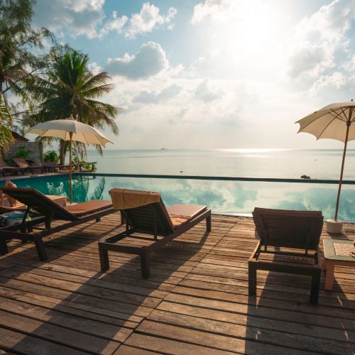 Lounge chairs and umbrellas by an infinity pool overlook a serene ocean view with palm trees under a bright sunlit sky.