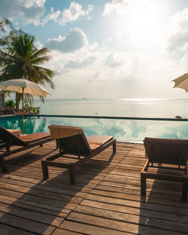 Lounge chairs and umbrellas by an infinity pool overlook a serene ocean view with palm trees under a bright sunlit sky.