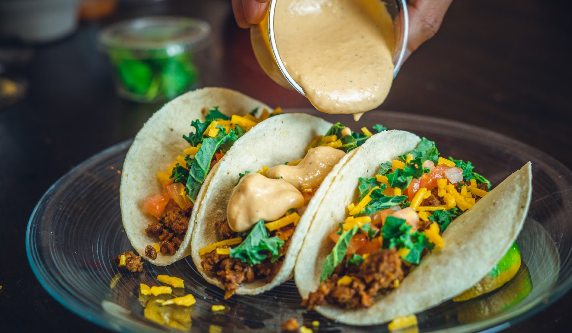 Three tacos with toppings like cheese, ground meat, and greens are being drizzled with a creamy sauce on a plate.