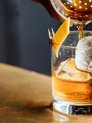A cocktail being poured into a glass with ice and a lemon twist garnish, set on a table.