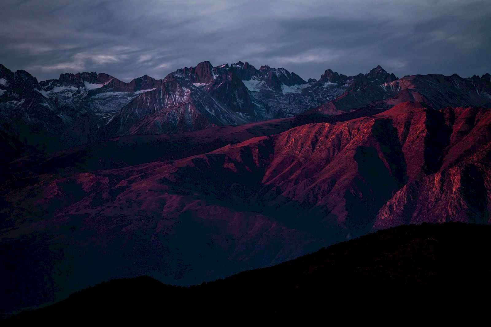 A mountain range is bathed in deep red sunlight under an overcast sky, creating a dramatic and atmospheric landscape scene.