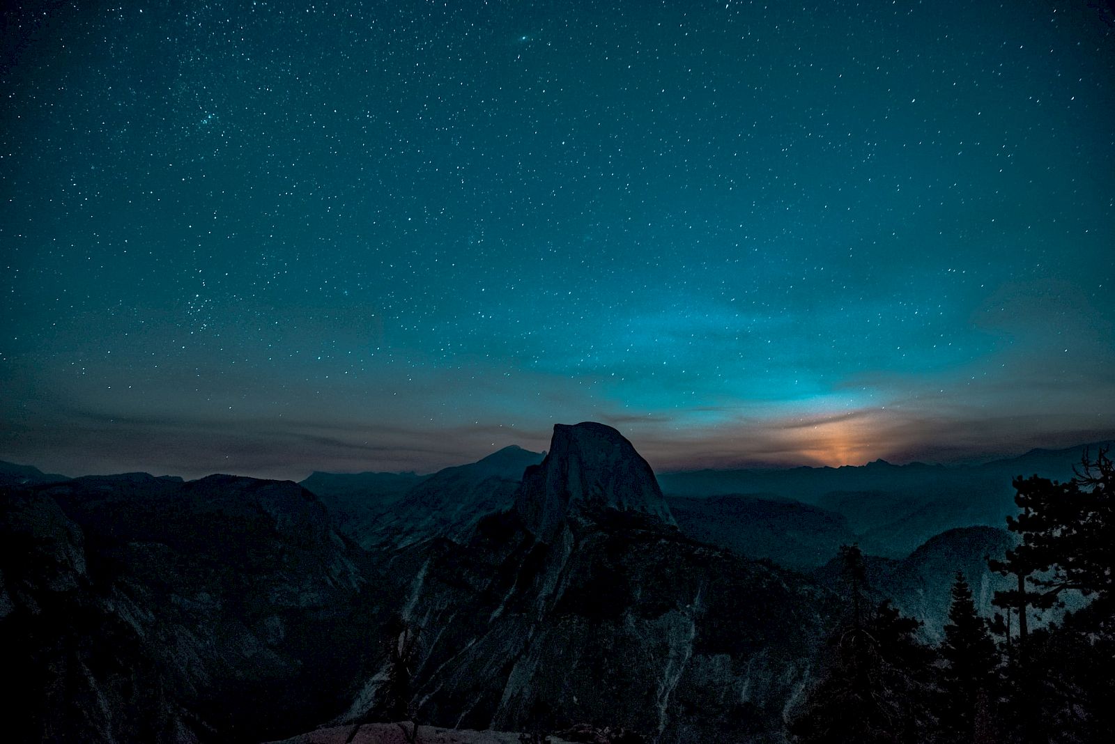 A starry night sky over a mountainous landscape, with a prominent peak silhouetted against the twilight horizon.