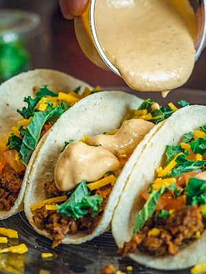 Three tacos with ground meat, cheese, lettuce, and tomatoes are topped with a creamy sauce being poured over them.