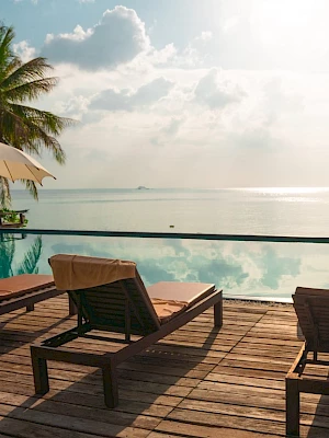 Poolside deck with lounge chairs and umbrellas, overlooking a serene ocean view with palm trees and cloudy sky in the background.