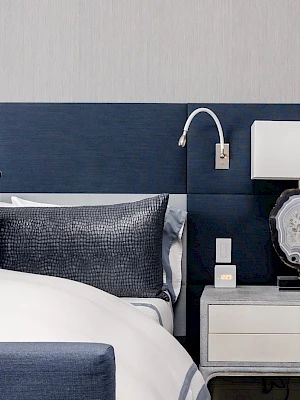 A modern bedroom features a bed with navy and white bedding, a blue headboard, a bedside table, and a stylish lamp.