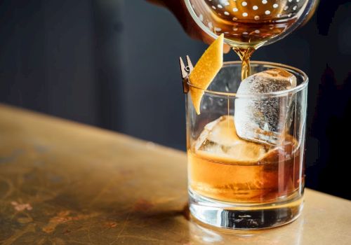 A glass of cocktail with ice and a lemon twist garnishing is being poured into on a bar counter.