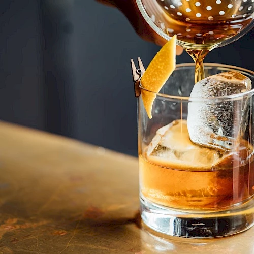 A glass of cocktail with ice and a lemon twist garnishing is being poured into on a bar counter.