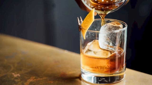 A glass of cocktail with ice and a lemon twist garnishing is being poured into on a bar counter.
