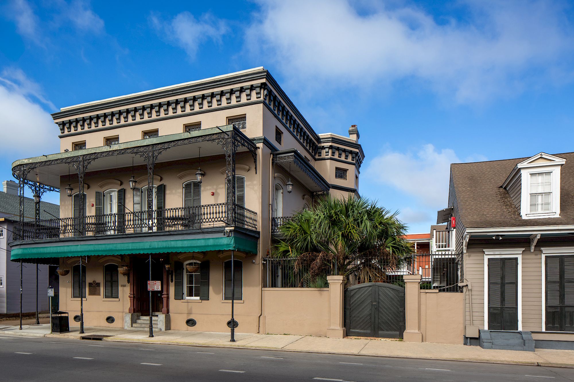 The image shows a historic building with wrought iron balconies and adjacent structures on a sunny day, likely in an urban area.