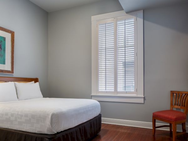 A simple room with a bed, chair, window, and wall art in neutral tones and a wooden floor.