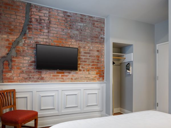 A room with a brick accent wall, mounted TV, wooden chair, and a partially visible bed, featuring a closet with hangers to the right.