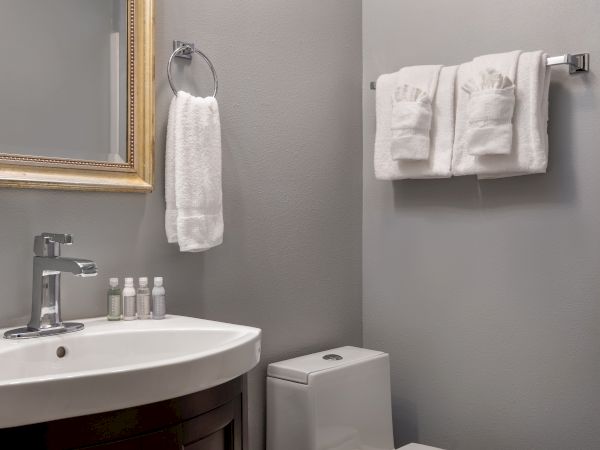 A modern bathroom with a sink, a mirror, toiletries, white towels on the rack, and a toilet against gray walls.
