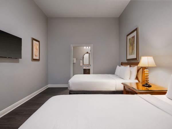 A hotel room with two beds, a TV on the wall, a wooden side table with a lamp, and framed art on gray walls.