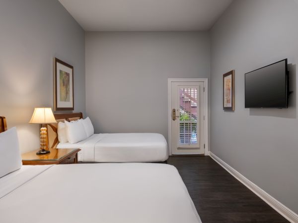 The image shows a hotel room with two beds, a nightstand with a lamp, wall art, a flat-screen TV, and a door leading outside.