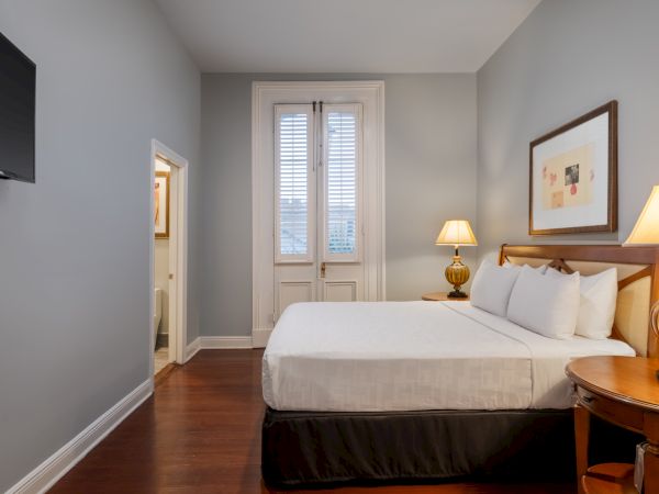The image shows a cozy bedroom with a neatly made bed, lamps, framed artwork, wooden flooring, and a wall-mounted TV.