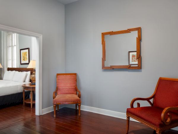 A room with two wooden chairs and a decorative frame on the wall, leading to a bedroom with a bed and nightstand visible.