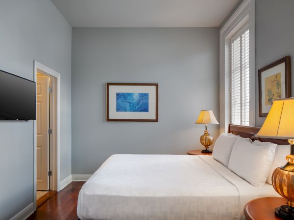 A cozy bedroom with a bed, two lamps, wall art, and a TV. Walls are light gray, enhancing the calm ambiance of the room.