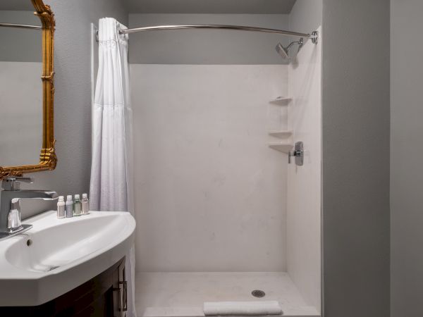 The image shows a bathroom with a shower, white curtain, sink, a mirror, and toiletries on the sink counter.