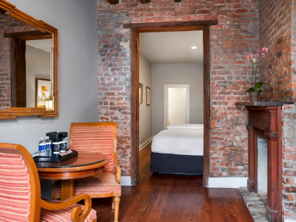 A cozy room with brick walls, a small table with chairs, a mirror, and a view into a bedroom with a bed and fireplace.