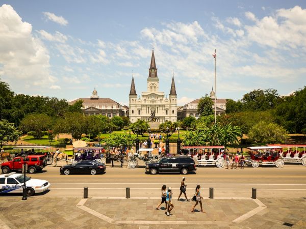 The image shows a crowded street with carriages in front of a historic building and park on a sunny day.