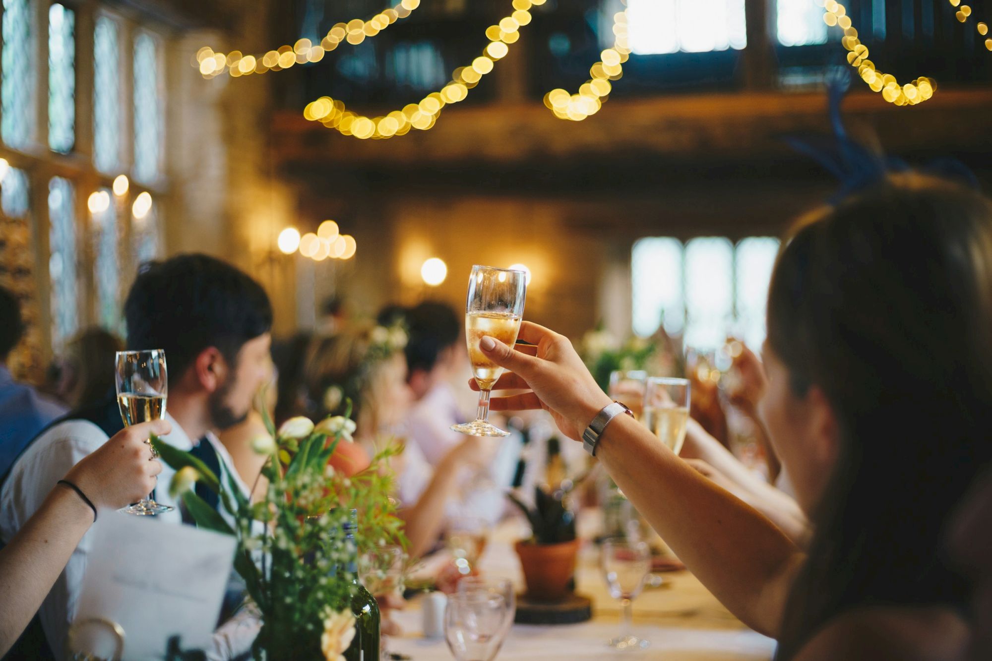 People are at a celebration, raising glasses for a toast in a warmly lit venue with decorations and flowers on tables.