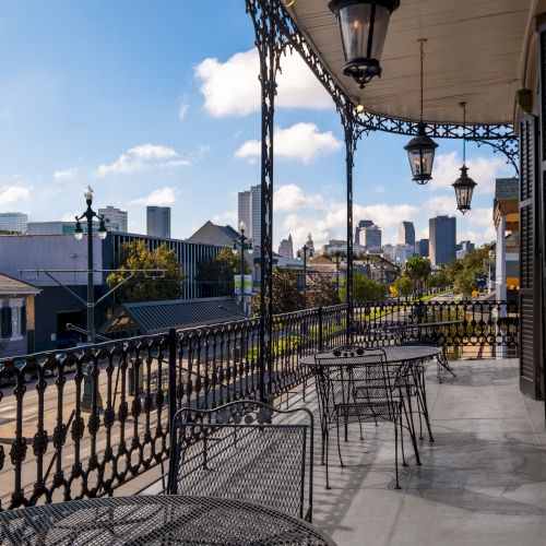 A balcony with wrought iron railings overlooking a street and skyline on a sunny day, with several metal tables and chairs arranged.