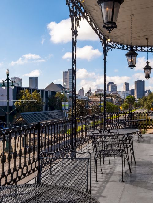 A balcony with wrought iron railings overlooking a street and skyline on a sunny day, with several metal tables and chairs arranged.