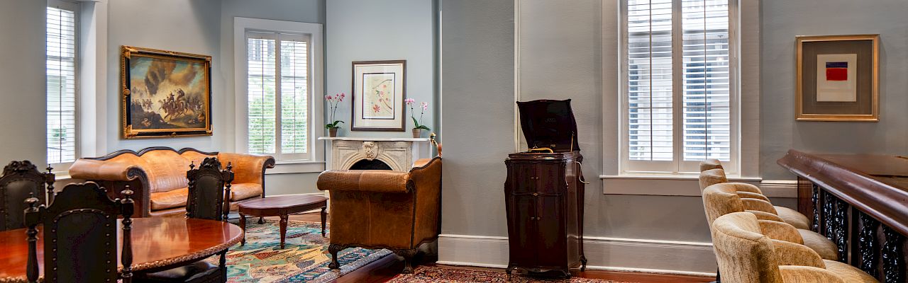 A spacious room with elegant furniture, patterned rugs, paintings, and a bar area with stools next to shuttered windows.