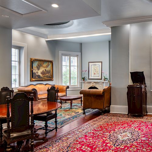 A spacious room with elegant furniture, patterned rugs, paintings, and a bar area with stools next to shuttered windows.
