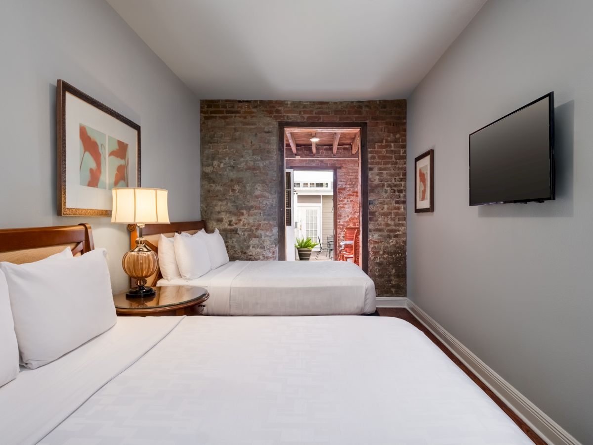 Two beds in a cozy room with a brick wall, wall-mounted TV, and artwork; a hallway or doorway leads outside, revealing greenery.