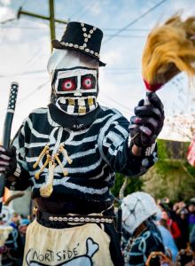 A person in a skeleton costume participates in a lively street parade, with a crowd and buildings in the background.