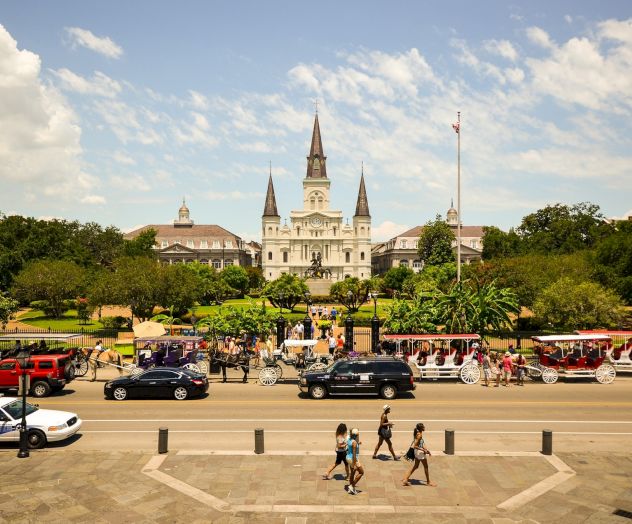 A historic cathedral with spires stands in a park setting, bustling with people, horse carriages, and cars under a blue sky.