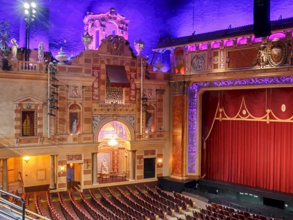 An ornate theater interior with balconies, red seating, and a grand stage with a red curtain, illuminated by colorful lights.