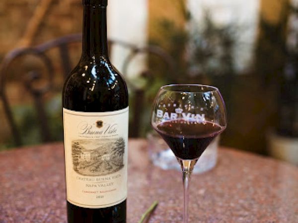 A wine bottle, filled glass, red rose, and a blue napkin on a table in a cozy setting.