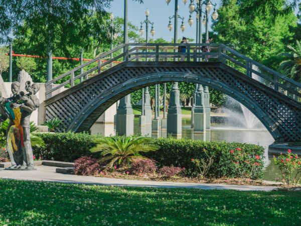 A scenic park features a wooden bridge over a pond, a fountain, green plants, and a modern sculpture amidst lush greenery and trees.