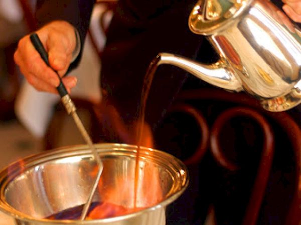 A person is preparing a flaming beverage by pouring liquid from a silver teapot into a bowl with fire at a restaurant table.